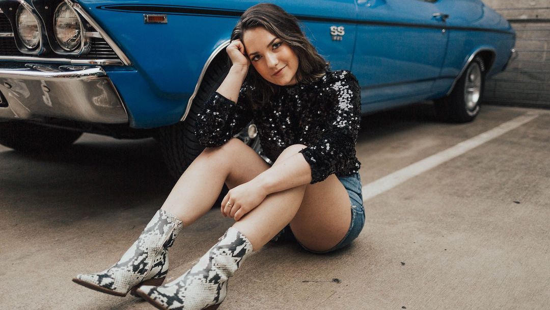 Country solo artist Haley Mae Campbell poses for a photo, seated on the ground in a parking lot next to an old Chevrolet SS